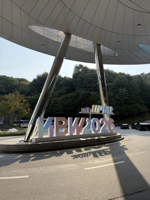 An outdoor signage installation for KBW2026 (Korea Blockchain Week) with 'with U-Pbit' text, featuring metallic letters beneath a modern architectural canopy.