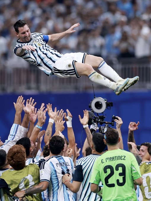 Lionel Messi in Argentina's white and sky-blue striped kit celebrating a World Cup victory by leaping into the air, being tossed by teammates including goalkeeper E. Martinez (#23) in a green jersey.
