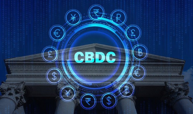 A classical bank building facade is overlaid with a glowing blue digital interface featuring the text "CBDC" at the center, surrounded by a circular arrangement of currency symbols including the dollar ($), euro (€), pound (£), rupee (₹), bitcoin (₿), and others against a backdrop of binary code. The image visually represents the central bank digital currency concept that the accompanying post addresses, illustrating the intersection of traditional banking architecture with digital currency technology that the Federal Reserve is now blocked from issuing until 2030.