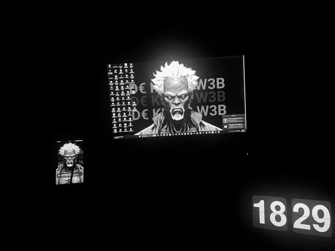 A dark room showing a computer monitor and phone displaying a black and white character wallpaper, with a digital clock reading '18 29' in the corner.