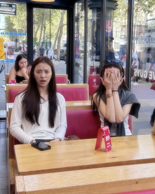 Two young women sit at a wooden table in a cafe or restaurant with large windows; the woman on the left speaks while the woman on the right covers her face with her hands, with a red table number '89' in front of them.