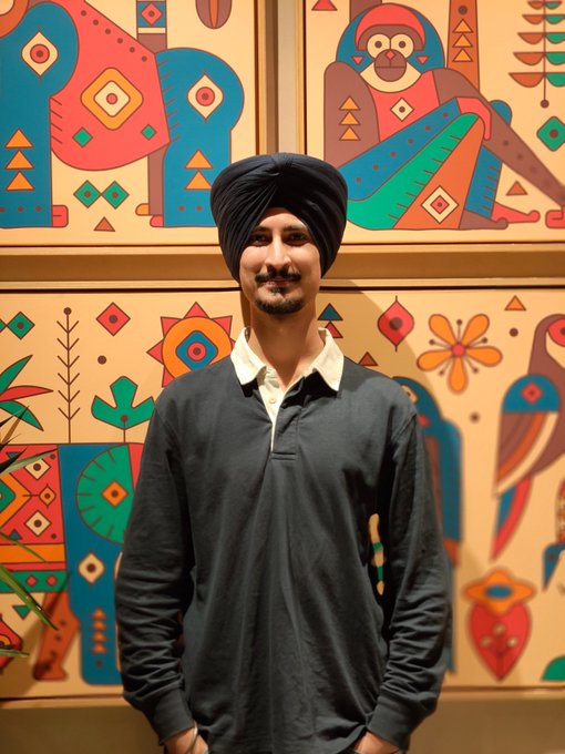 A man wearing a black turban and dark long-sleeve shirt with a white collar stands smiling in front of colorful, geometric tribal-style artwork featuring stylized animals and patterns, appearing to be at the Logos Network event mentioned in the post.