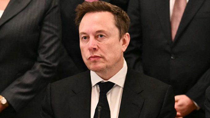 Elon Musk, wearing a black suit and tie, is photographed in a formal setting.