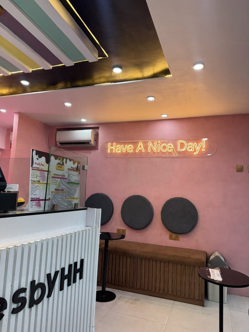 The interior of a pink-walled shop features a neon sign reading "Have A Nice Day!!" above a seating area with black circular cushions on the wall and a counter labeled "...ssbyHH".