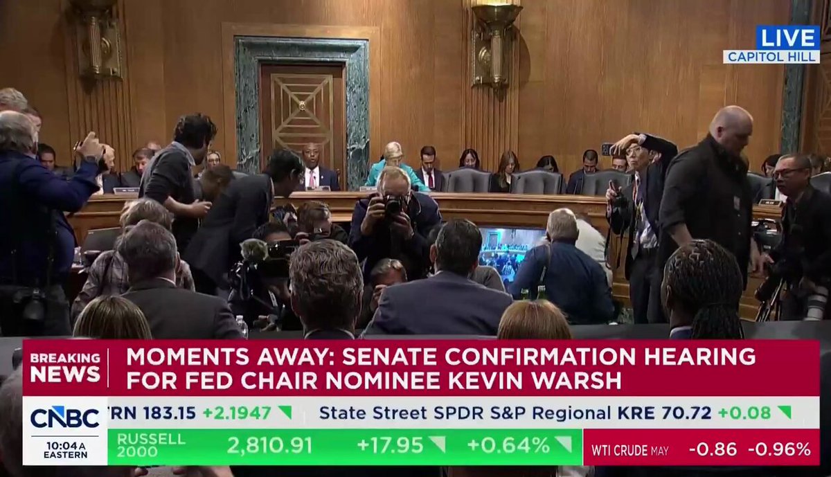 The image shows a news broadcast of a Senate hearing, with people sitting in chairs around a long table as they listen to the confirmation hearing for Fed Chair Kevin Warsh. The news anchor is reporting that the confirmation hearing has officially begun, and the first Federal Reserve Chair has been confirmed.
