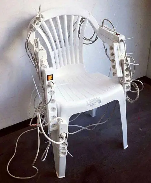 A white plastic armchair with numerous power strips, extension cords, and adapters tangled and mounted all around its frame, plugged into multiple outlets.