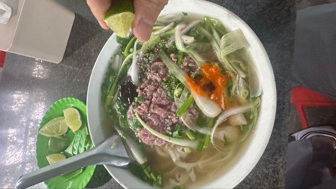 The image shows a bowl of soup with various ingredients such as noodles, meat, onions, and peppers. A person's hand is reaching into the bowl to add some lime juice or lemon juice over the top of the soup. The scene also includes other bowls on the table, suggesting that this could be a restaurant setting where people can enjoy different types of dishes.