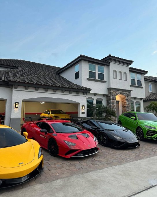 The image features a row of luxury sports cars parked outside a large white house, including a red Lamborghini, a yellow Porsche, and other vehicles. The cars are positioned next to each other, showcasing their sleek designs and vibrant colors. The scene suggests that these cars belong to someone who takes pride in their collection and possibly has an interest in the automotive industry or luxury lifestyle.