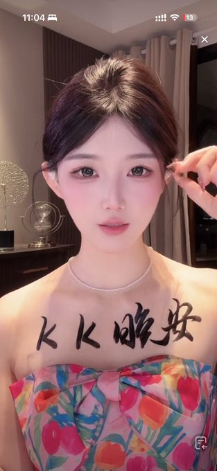 A selfie of a young woman with dark hair and large eyes, wearing a floral strapless top and pearl necklace, with Chinese characters written on her chest and a timestamp of 11:04 displayed at the top.
