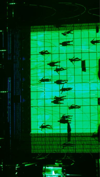 A stylized green-lit photograph of a vertical indoor skydiving wind tunnel, showing multiple silhouetted figures flying against the bright green airflow.