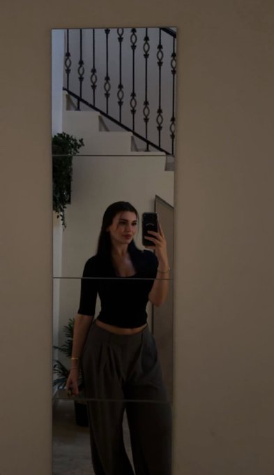 A mirror selfie of a woman wearing a black crop top and loose-fitting gray pants, taken in front of a mirror near a staircase with wrought iron railings.