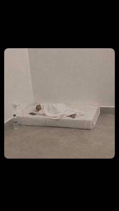 A minimalist room with concrete floors and white walls, showing a small mattress on the floor where a person lies covered in white blankets, with a phone charging nearby and a water bottle on the floor.