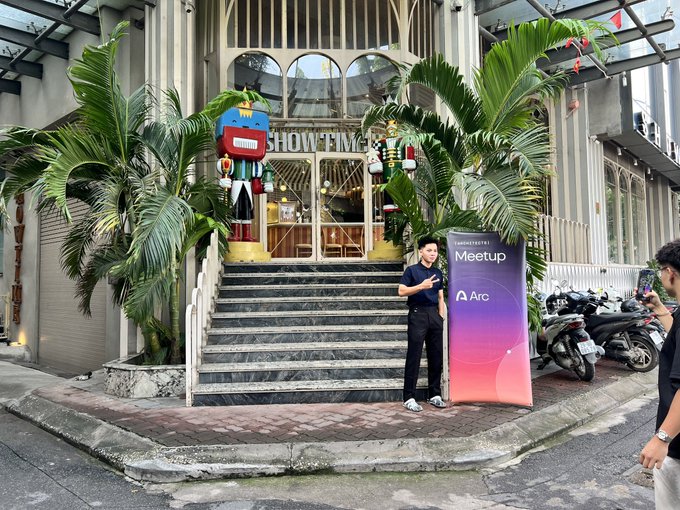 The user wants a concise caption describing the image.
I need to identify key elements:
- A man standing on a sidewalk next to a set of stairs.
- He is holding a microphone and standing next to a vertical banner.
- The banner says "TechCrunch Taipei Meetup" and has the logo "Arc".
- Behind him are large steps leading to a building entrance with "SHOWTIME" visible above the door.
- There are large nutcracker statues flanking the entrance.
- There are palm trees.
- To the right, another person is partially visible taking a photo or video.
- Motorcycles are parked in the background.
- The text on the banner is "TechCrunch Taipei Meetup" and "Arc".
- The prompt mentions a post saying "GM Chờ các @Arc chitect đến". This translates roughly to "GM Waiting for the @Arc architects to arrive". This context explains why the man is waiting/standing there. The man in the image looks like