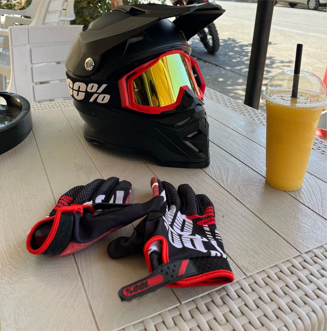 A black motorcycle helmet with "100%" branding and red-accented gloves sit on a tiled outdoor table alongside an iced orange drink, suggesting the rider is taking a break to recharge after a ride.
