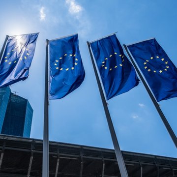 Four European Union flags fly against a bright blue sky, symbolizing the EU institutions pushing to amend the bloc's MiCA stablecoin regulations. The image visually reinforces the article's focus on ECB and EU central bank efforts to reshape crypto oversight.