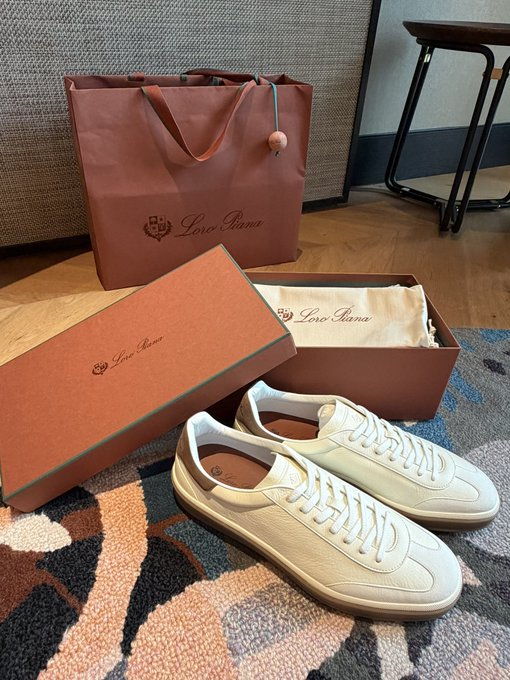 Image 9: A pair of white leather Loro Piana sneakers displayed next to their branded orange box and shopping bag, set on a patterned rug.