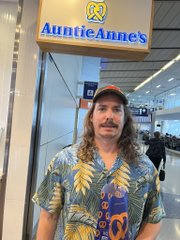 A man with long hair and a mustache wearing a tropical print shirt and a baseball cap, standing in front of the illuminated 'Auntie Anne's' sign at what appears to be a mall or commercial space.