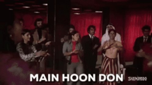 The image features a group of people standing together, with one person holding up a sign that says "Main Hoon Don" above their head. The background includes a red curtain and a man wearing a tie. There are also some text elements in the image, such as the words "SPY" and "puts", which could be related to the post or the event taking place.