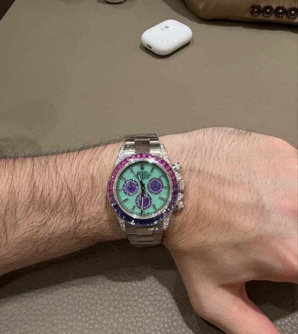 A wrist displays a Rolex watch featuring a diamond-encrusted bezel with pink and blue gemstones and a light green dial with purple sub-dials, photographed near a pair of white wireless earbuds.