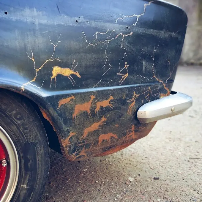 A close-up of a vintage vehicle's rear fender showing extensive rust and weathering, decorated with primitive-style rock art paintings of deer and human figures in orange/gold tones against the dark, cracked paint.