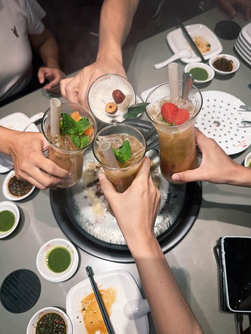 Multiple hands clink tall glasses filled with iced drinks garnished with mint, strawberries, and chocolate in a celebratory toast over a table laden with plates and dipping sauces.