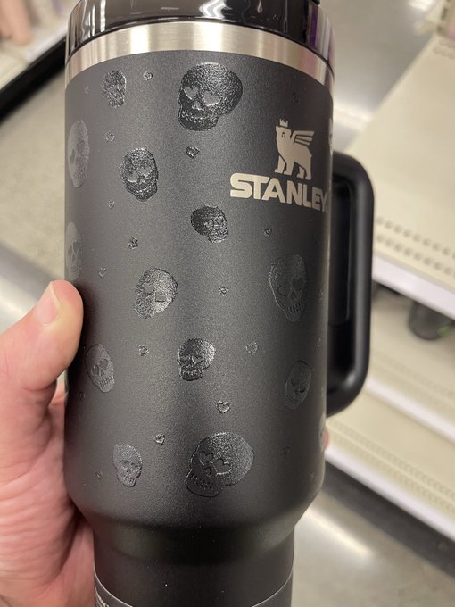 A hand holding a black Stanley thermos featuring skull patterns and the brand's lion logo, with what appears to be the moon's surface visible in the background.