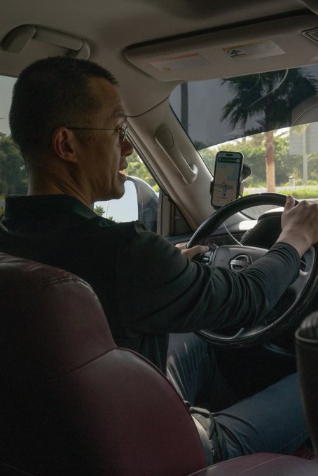 A man is driving a Nissan vehicle while a smartphone mounted on the dashboard displays a navigation map. The image does not visually depict any Binance or OKX branding or logos, nor does it show identifiable individuals associated with those companies, so the connection to the caption referencing CZ and Star figures remains unverified by the visual content.