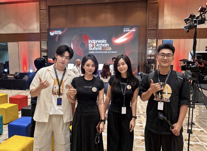 Four attendees pose in matching black ASTER t-shirts and lanyards in front of a stage displaying "Indonesia Blockchain Summit 2026 - Turning Infrastructure Into Impact," with a man on the left and a man on the right flashing hand gestures, ready for the upcoming trading competition at the main stage.