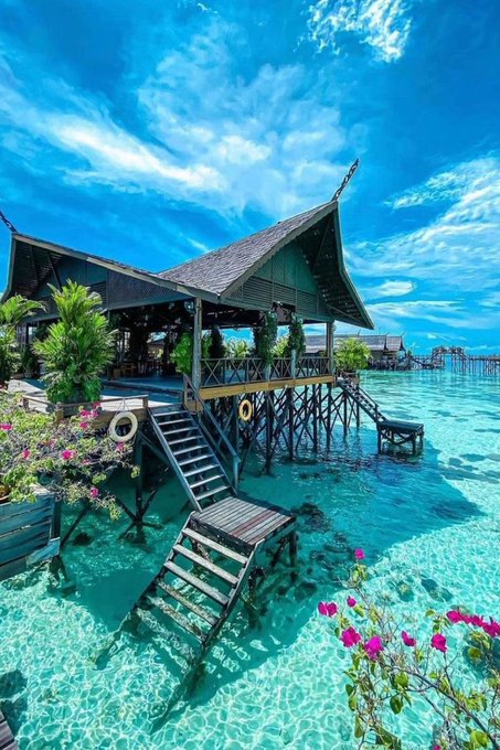 Scenic photograph of traditional wooden stilt houses built over crystal-clear turquoise water under a bright blue sky with scattered clouds, with pink flowers in the foreground.