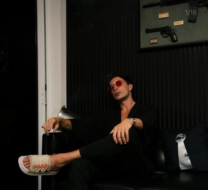 A man with red-tinted round sunglasses lounges on a black leather couch in a dimly lit room, holding a cigarette while wearing a relaxed black outfit and white slide sandals, with a display of firearms and ammunition mounted on the dark paneled wall behind him. The "1/16" in the corner indicates this is the first image in a series accompanying allegations against Luke Belmar, where the subject's seemingly cool, confident demeanor visually reinforces the post's claims of him being deceptive and tied to controversies.