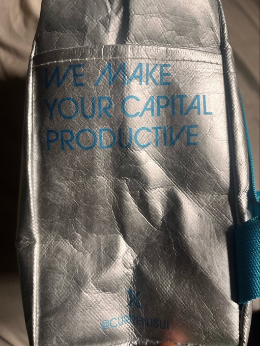 A silver textured fabric item, possibly a bag or pouch, displays the text 'WE MAKE YOUR CAPITAL PRODUCTIVE' in blue lettering, with '@CURRENTSU' partially visible below.