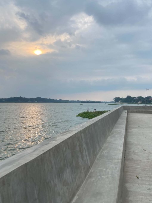 A waterfront view at dusk or dawn showing a concrete promenade and low sea wall along a calm body of water, with the sun glowing behind clouds, distant tree-lined shores, and a small patch of vegetation where two birds are perched.