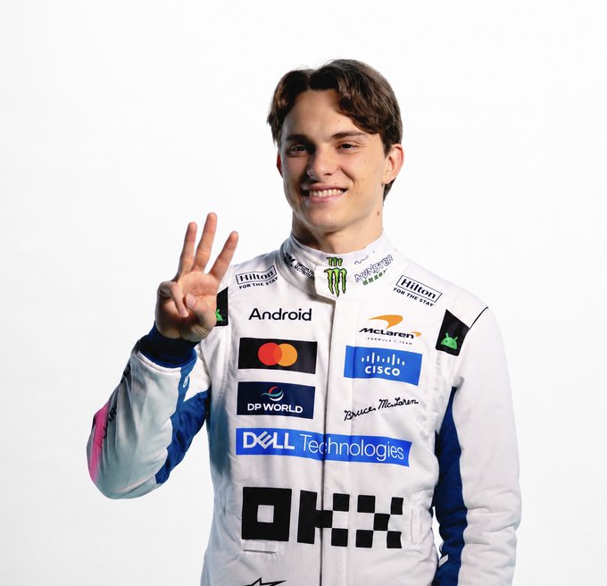 The image shows a young McLaren Formula 1 driver in a team racing suit featuring sponsors like OKX, Android, Dell Technologies, Cisco, and Mastercard, smiling and holding up three fingers. The pose connects to the quiz about OKX's European licenses, visually hinting at the number-related answer to the question.