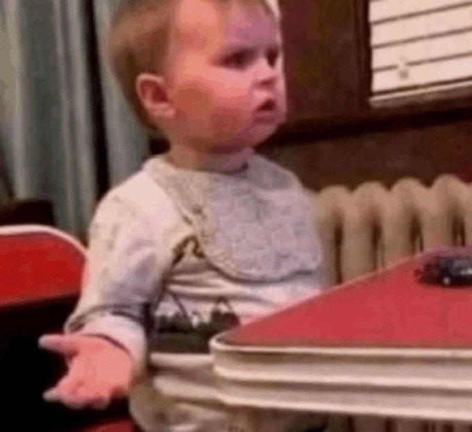 A well-known internet meme image of a baby (Hasbulla) in a bib looking confusedly at a red book on a table, from an old TV show clip.