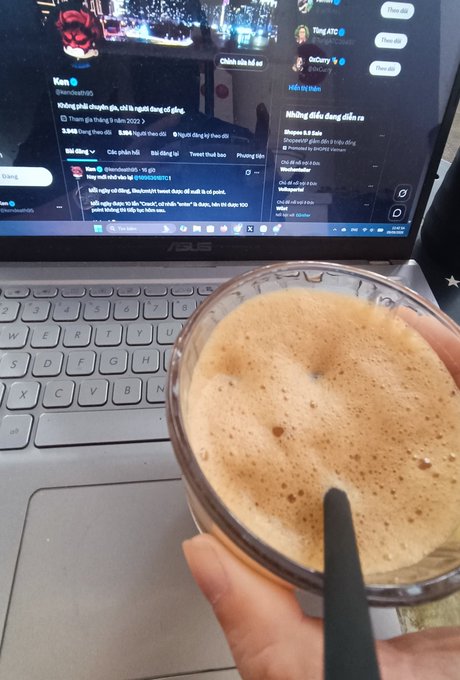 A hand holds a glass of frothy, light brown coffee over an ASUS laptop keyboard, with a social media feed displaying Vietnamese text visible on the screen behind it.