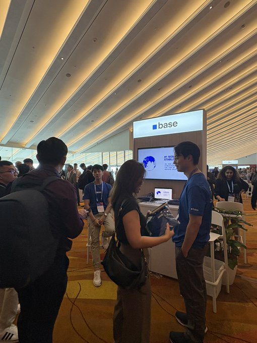 A group of people gather around a "base" booth featuring a screen displaying "#1 ROBOT STABLE" at what appears to be a tech or crypto conference, with attendees engaging in discussions about blockchain services.
