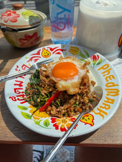 A photograph of a Thai dish (pad kra pao) served on a decorative Phed Mark branded plate, topped with a fried egg, accompanied by a bottle of water.
