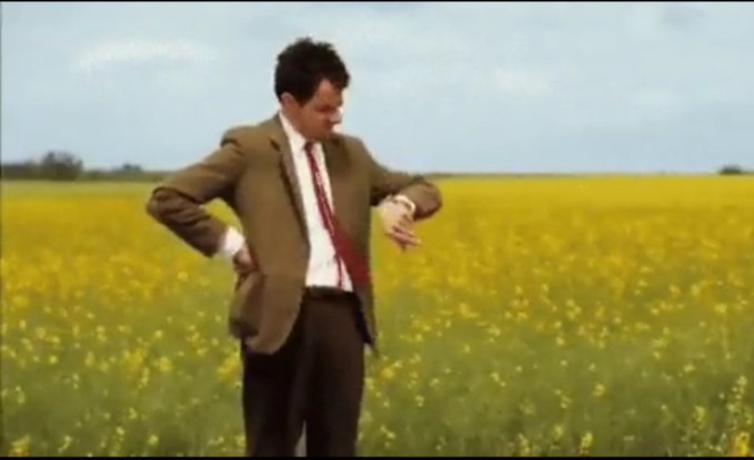 The image shows Mr. Bean (Rowan Atkinson), dressed in his iconic brown suit and red tie, standing in a vast yellow field looking impatiently at his wristwatch. The caption relates to the post as a humorous metaphor—Mr. Bean impatiently waiting in an endless field mirrors the creator's hopeful anticipation for their monetization payout to arrive.
