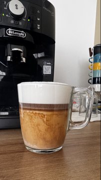 A glass mug filled with layered espresso and milk sits on a wooden surface in front of a black DeLonghi espresso machine.