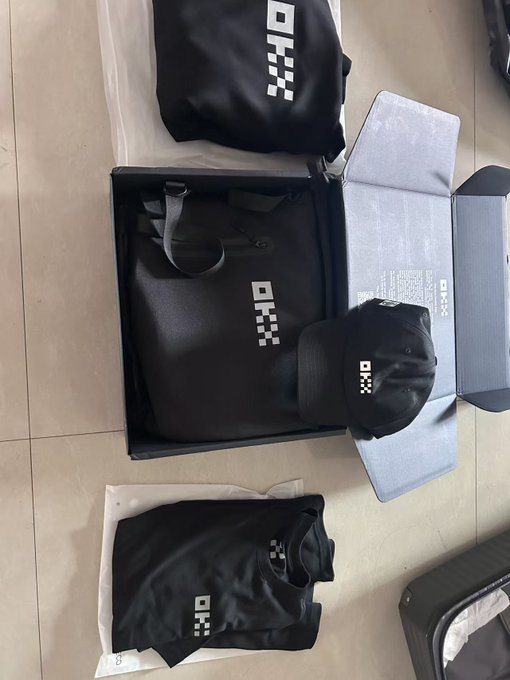 An overhead view shows newly opened OKX merchandise laid out on a tiled floor, including a black backpack and cap featuring the OKX pixelated logo, plus a t-shirt and hoodie in their packaging. The arrangement reflects the unboxing of the OKX swag haul mentioned in the post, with all items displaying the brand's signature black-and-white logo design.
