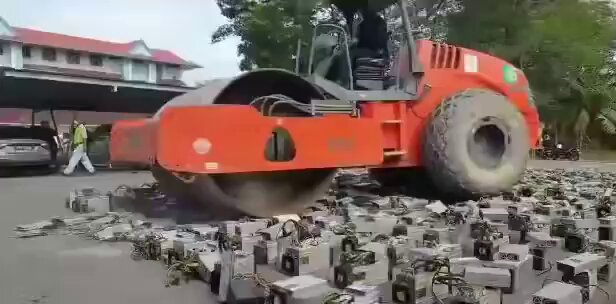 The image shows an orange construction vehicle, possibly a bulldozer or a pneumatic drill, lying flat on its side and surrounded by rubble and debris. The vehicles' position suggests that they have been crushed by some force or machinery. There are multiple objects scattered around in the scene, including some small boxes and bags of hardware, indicating recent activity or construction work nearby.