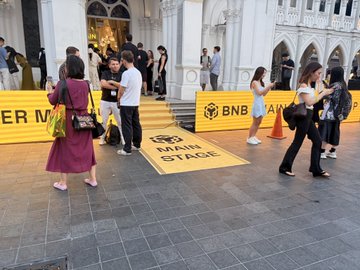 An outdoor gathering with people walking on a paved area featuring a 'BNB MAIN STAGE' logo on the ground, near a building with arched windows.
