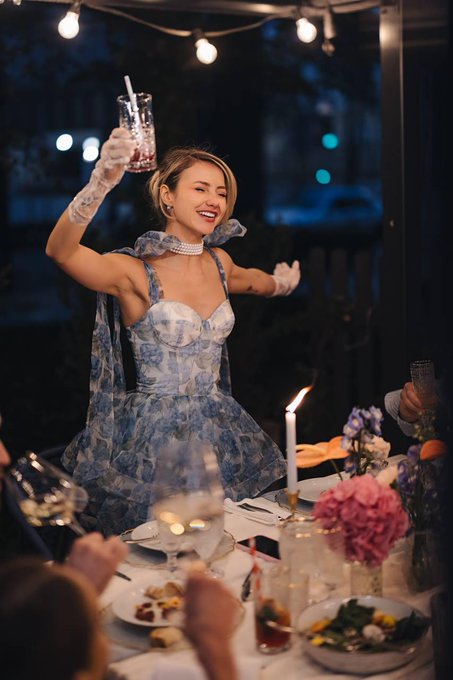 A woman in a floral blue and white dress with white gloves and pearl choker smiles joyfully while raising a drink in a toast at a candlelit dinner table.