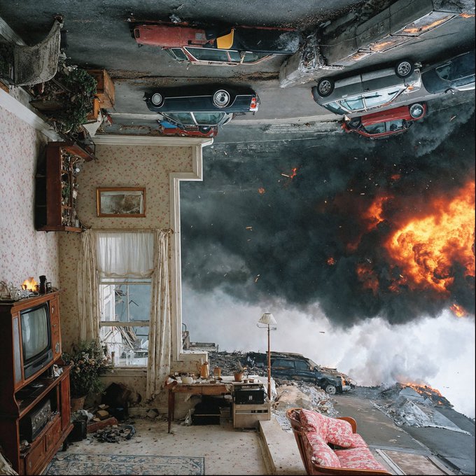 A surreal, upside-down scene where a vintage living room interior in the foreground contrasts with a chaotic street outside showing burning cars and an explosion.