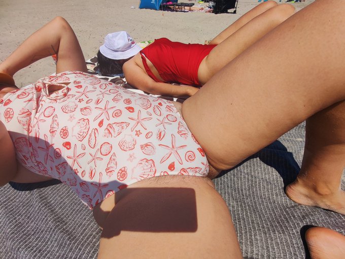 Two people sunbathe on a beach towel, one wearing a seashell and starfish patterned swimsuit and the other in a red swimsuit, with a white sun hat and other beachgoers visible in the background, capturing the carefree "sun out bush out" (girls out) summer vibe.