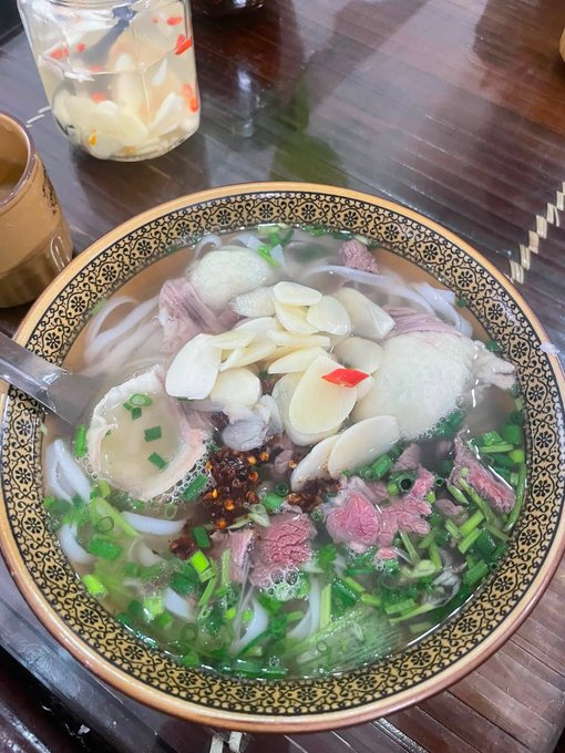 A top-down view of a large bowl of Vietnamese pho noodle soup with sliced beef, white onions, green herbs, and chili, accompanied by a side jar of pickled vegetables on a wooden table.