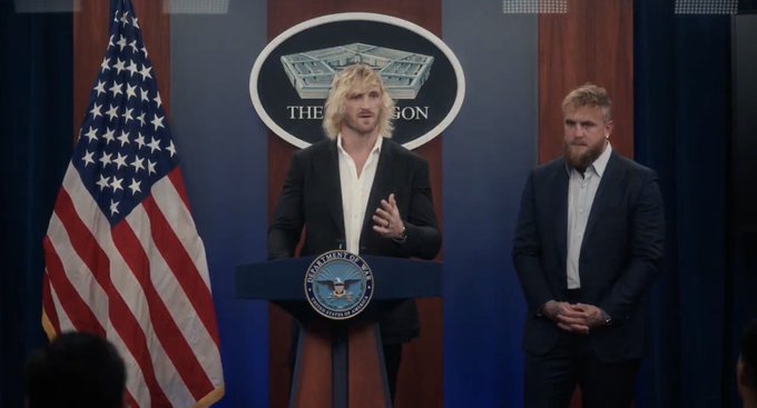 A man with blonde hair and a beard in a dark suit speaks at a podium bearing "The Pentagon" seal, with an American flag beside him and another bearded man in a suit standing to his right. The image appears to be from the comedy film "The Interview," aligning with the post's praise for the movie.