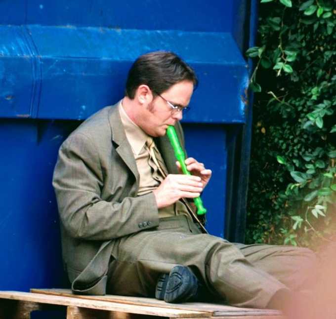 A man in a brown suit and tie with sideburns sits against a blue backdrop, playing a green recorder flute.