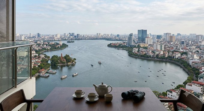 A scenic view from a balcony overlooking a river and city, featuring a table set with a teapot, cups, and a camera.