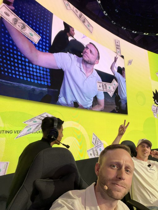 A man takes a selfie at an event, with a large screen behind him showing himself tossing dollar bills alongside other attendees; the backdrop features "Inviting Ve..." text and cartoon graphics. The image plays into the caption's playful "Not real (yet)" message, highlighting the staged, photo-ready moment before his upcoming Sui Basecamp appearance tomorrow.